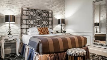 A bedroom with a white brick wall, a decorative headboard, a striped bedspread, a bench, two bedside tables with lamps, and a large mirror.