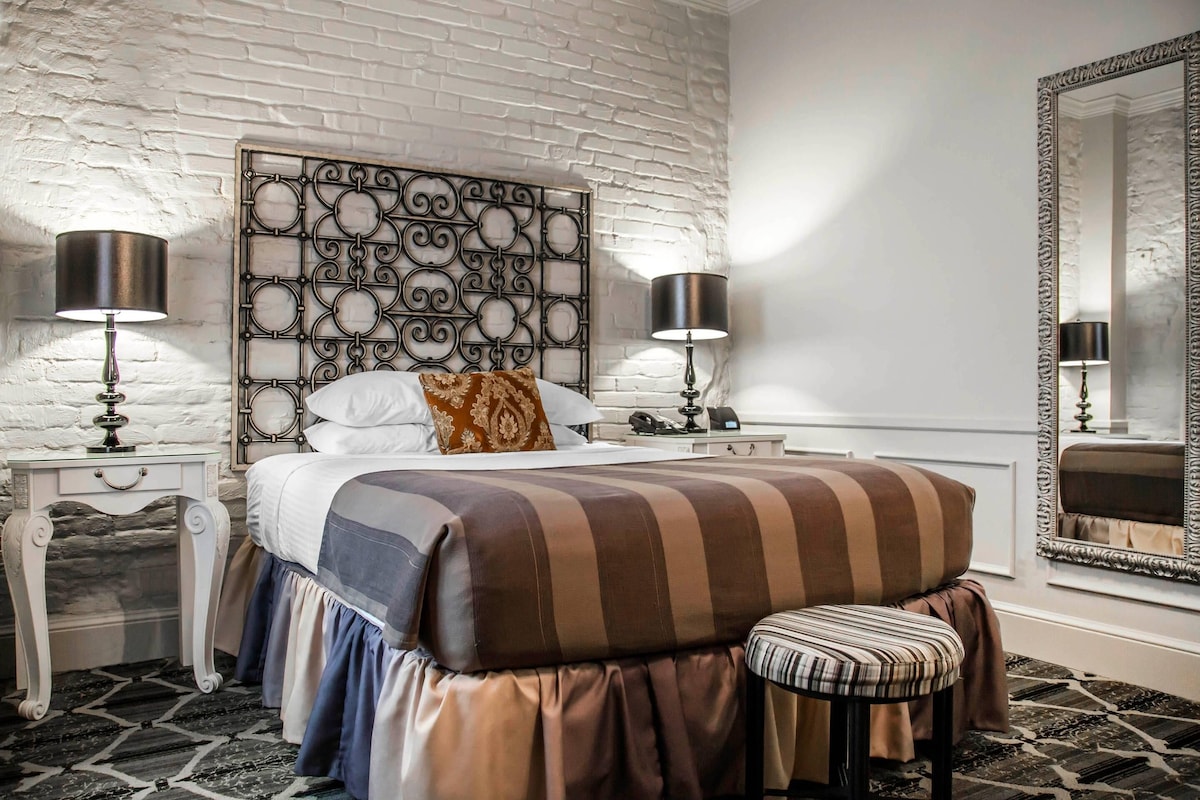 A bedroom with a patterned headboard, a bed with striped bedding, a bench, two bedside tables with lamps, and a large mirror.