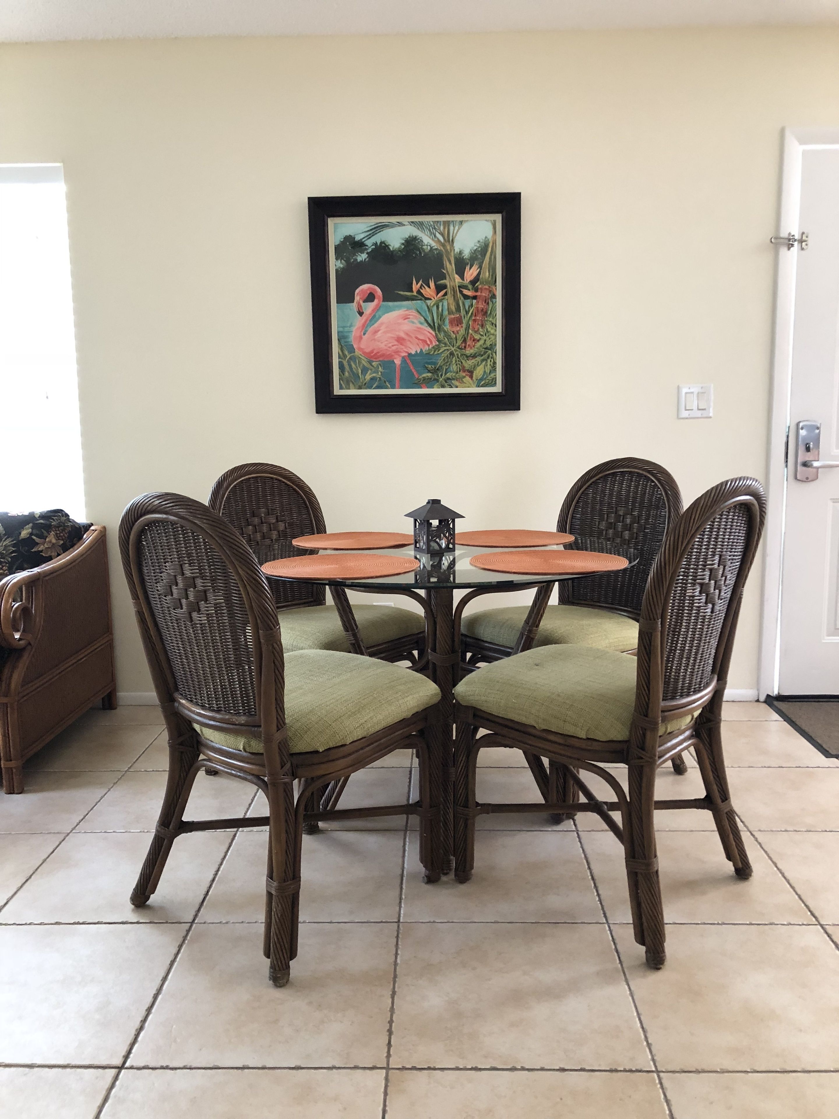 A dining area with a round table, four wicker chairs with green cushions, and a framed painting of a flamingo on the wall.