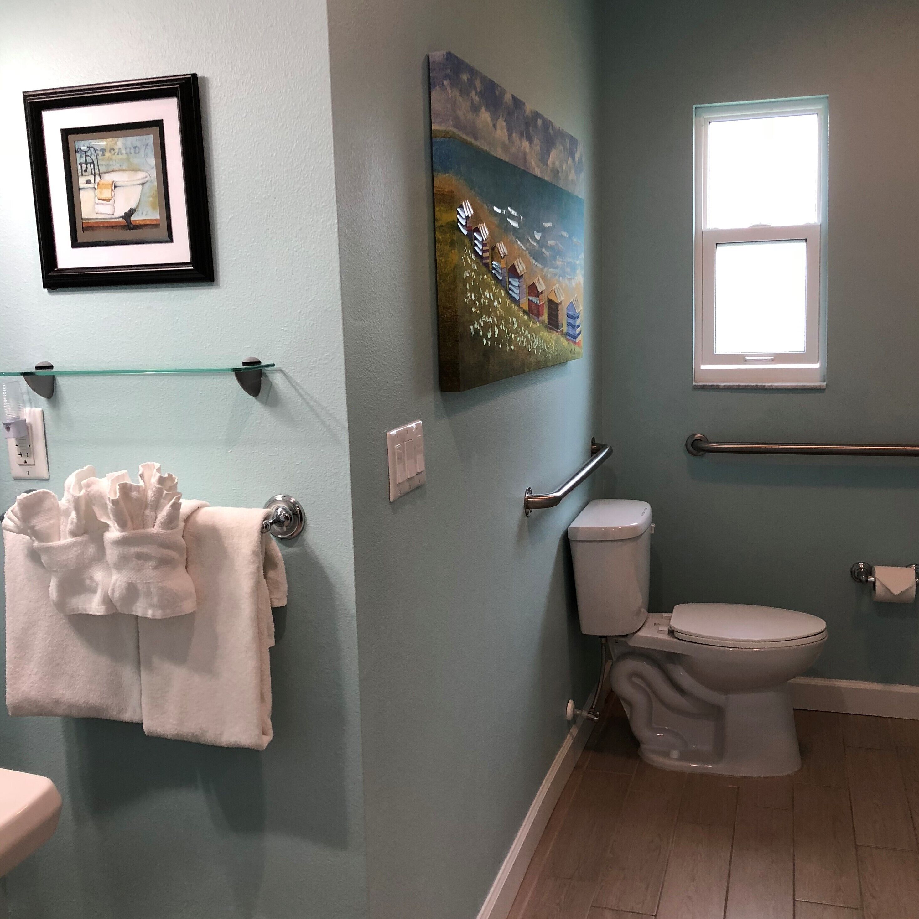 A bathroom with a toilet, a sink, a glass shelf with a towel, a painting, and a window.