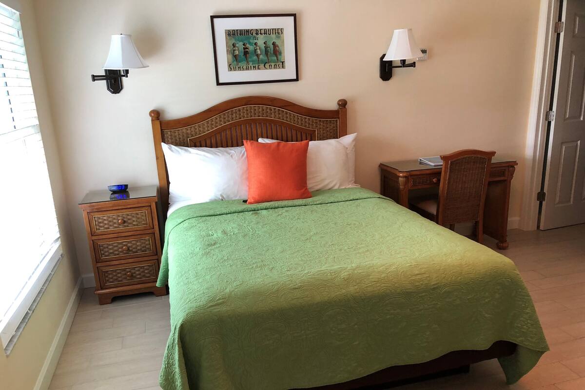 Een net opgemaakt bed met een groene sprei, een houten hoofdbord en twee nachtkastjes met lampen.
