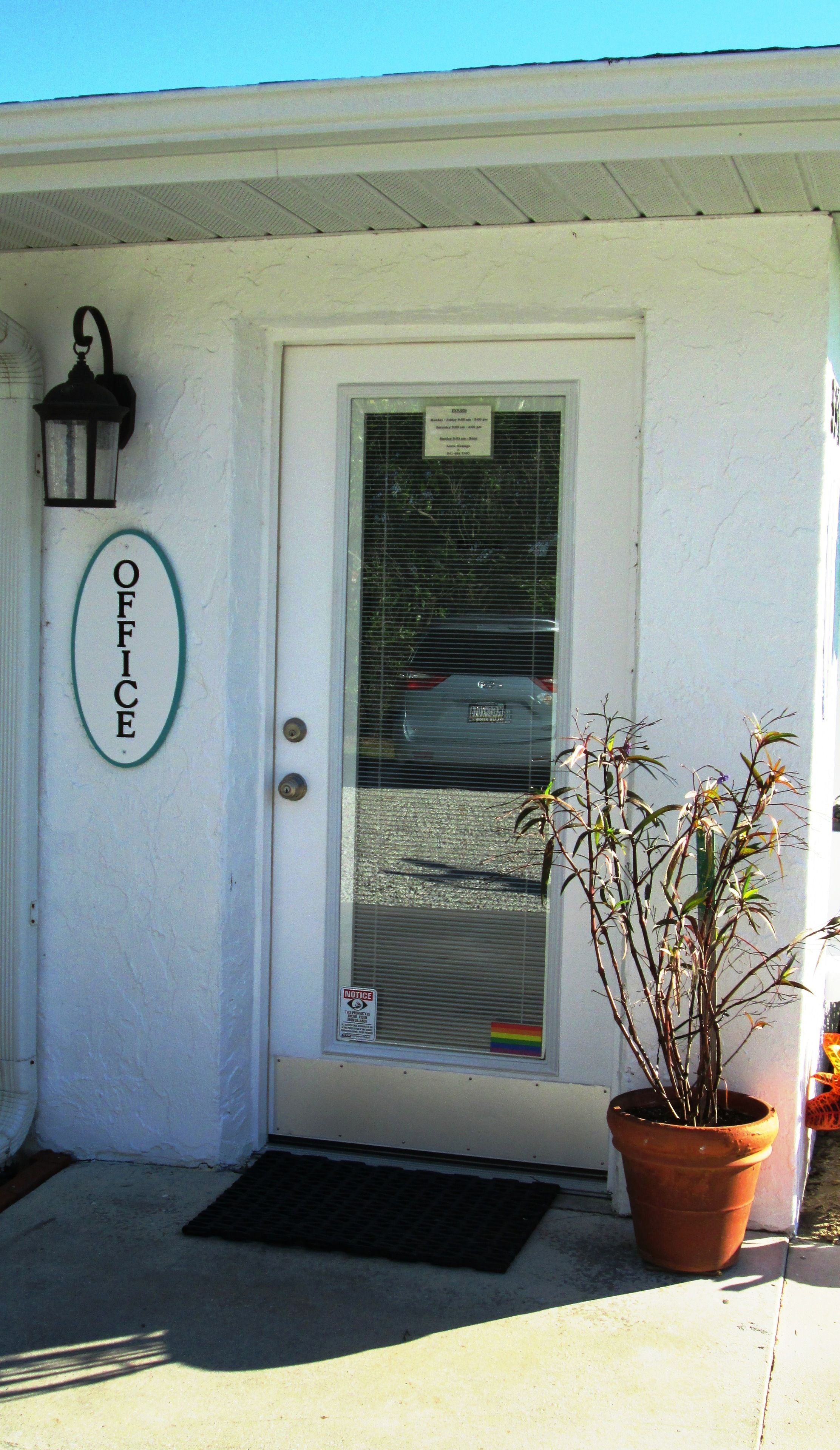 A white building with a door, a sign reading "OFFICE", a potted plant, and a black mat at the entrance.