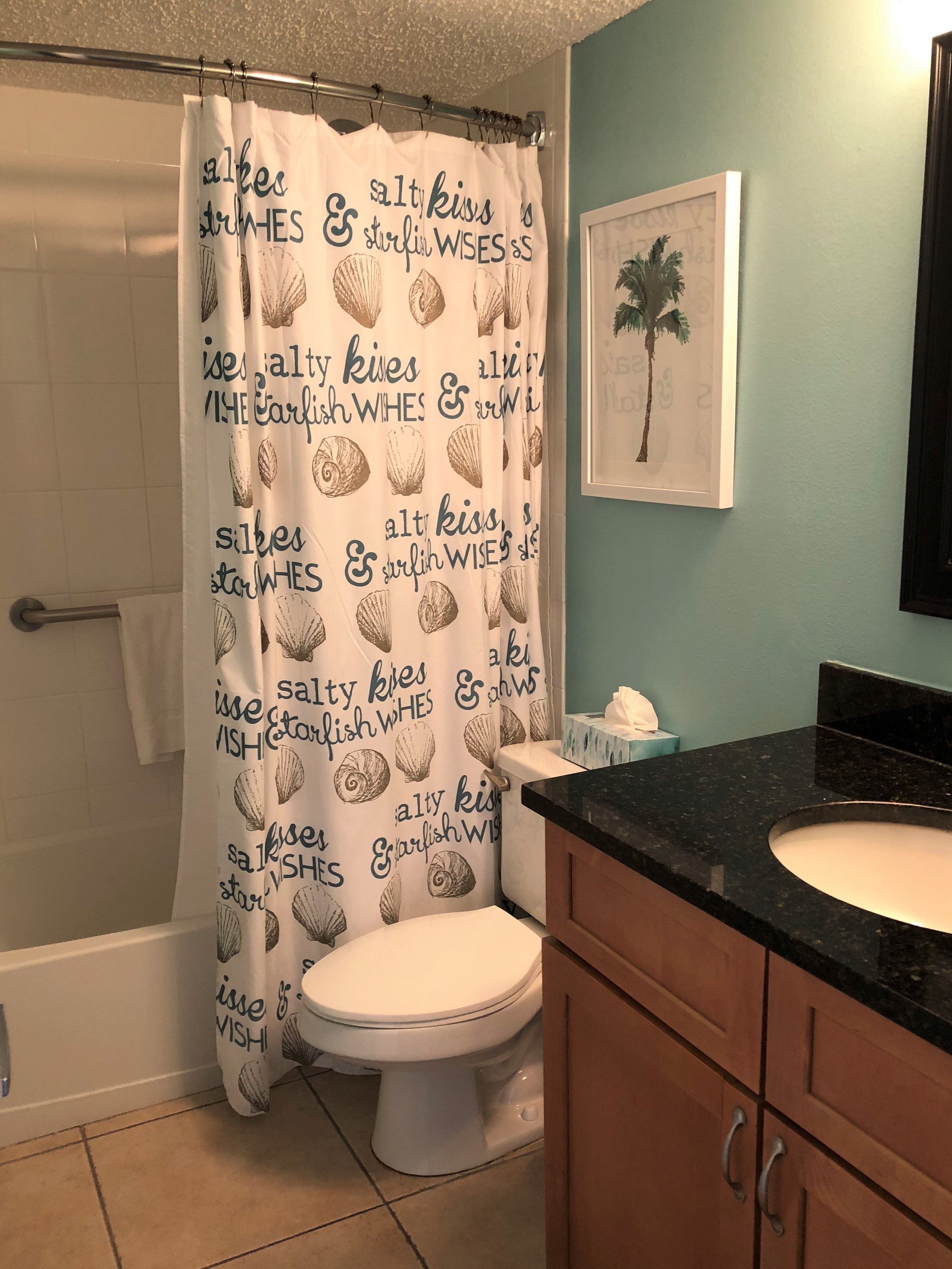 Une salle de bain avec un rideau de douche orné de motifs de coquillages et d’un texte, des toilettes, un lavabo et, au mur, un tableau représentant un palmier.