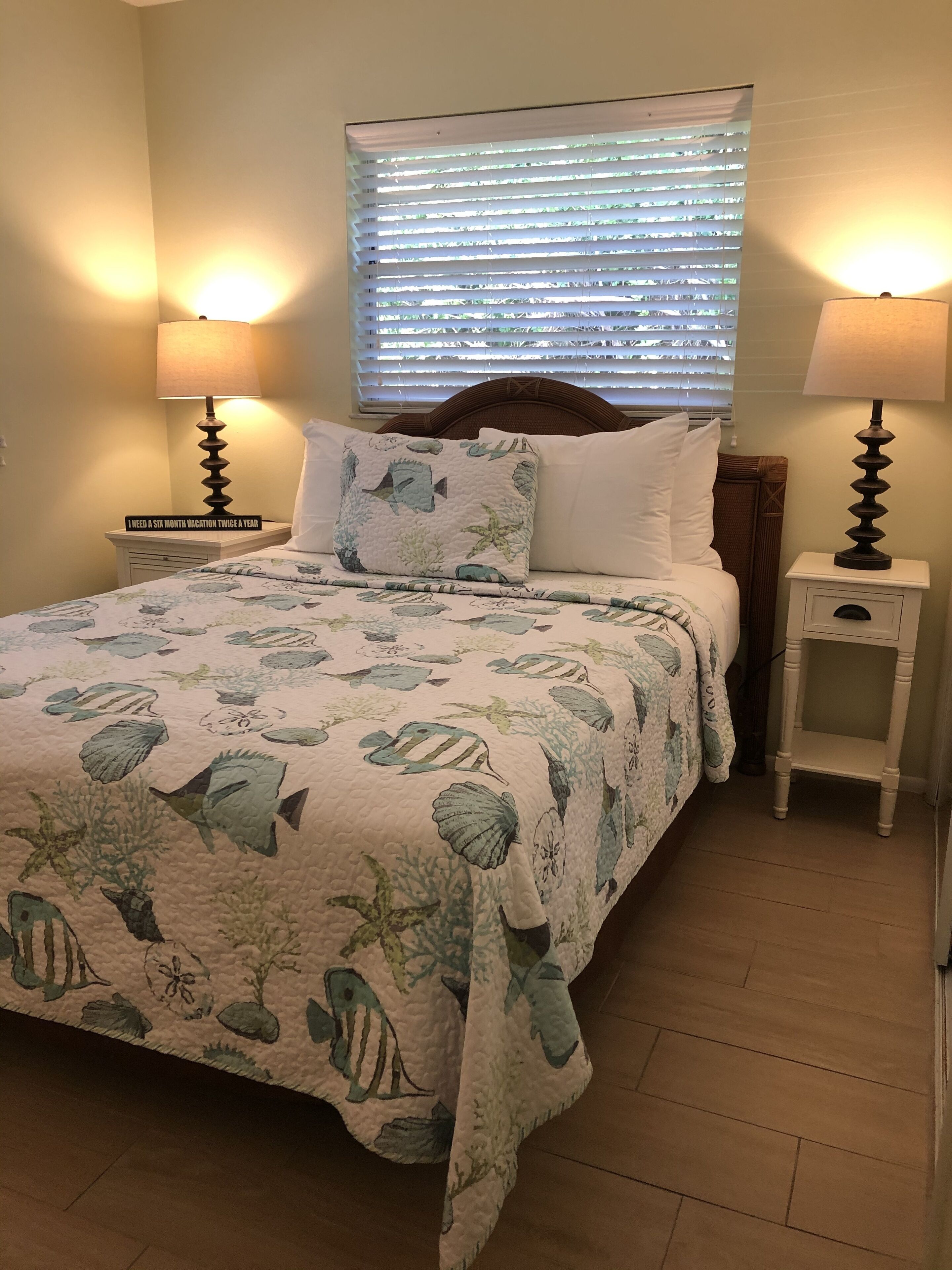 A bedroom with a bed, two bedside tables, a nightstand, a window with blinds, and a wall-mounted lamp.