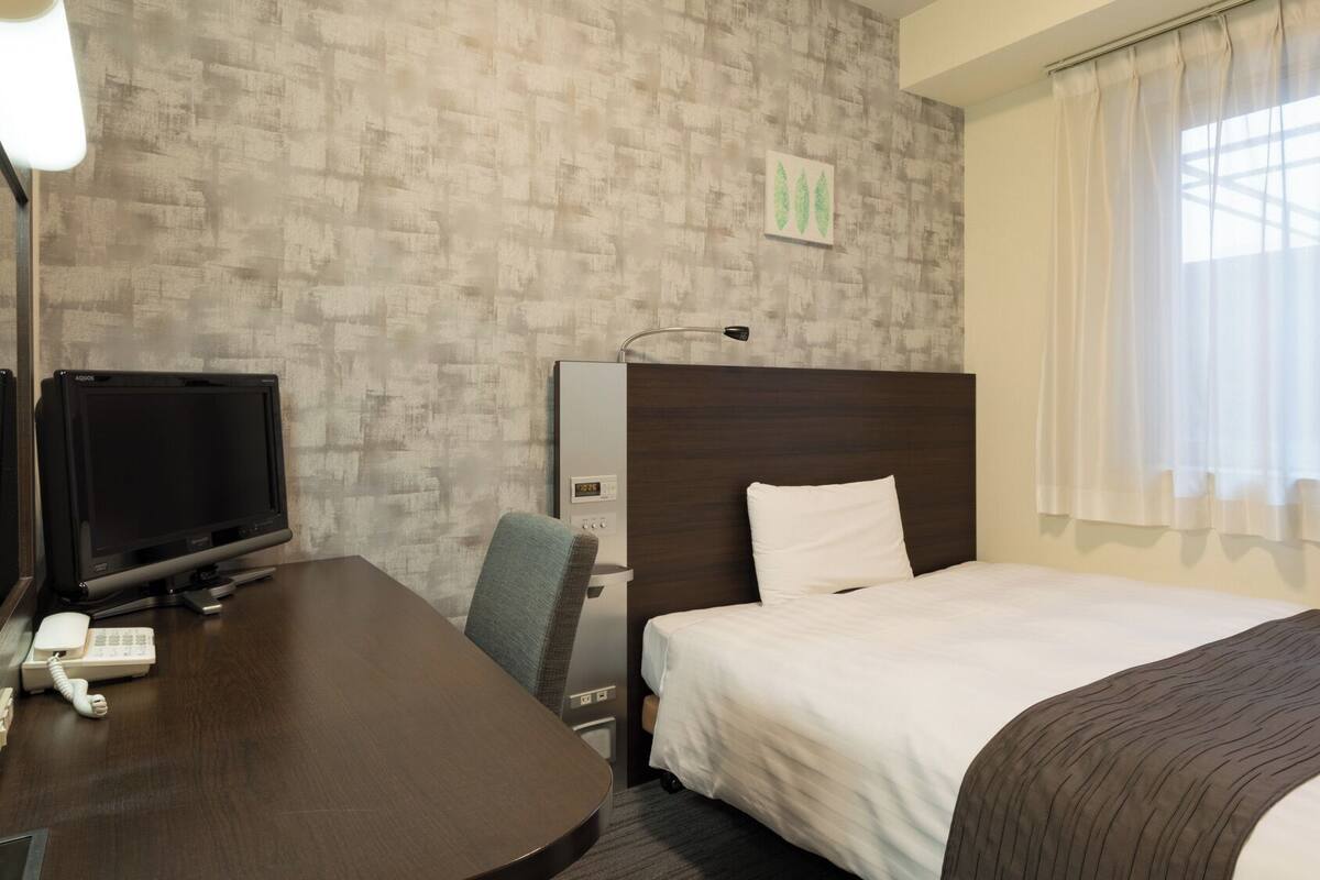 A hotel room with a bed, a desk with a television, a telephone, a headboard, and a window with curtains.