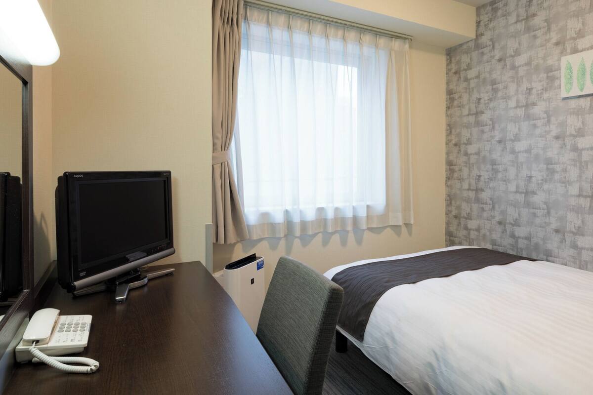 A hotel room with a bed, a desk with a television, a telephone, and a window with curtains.