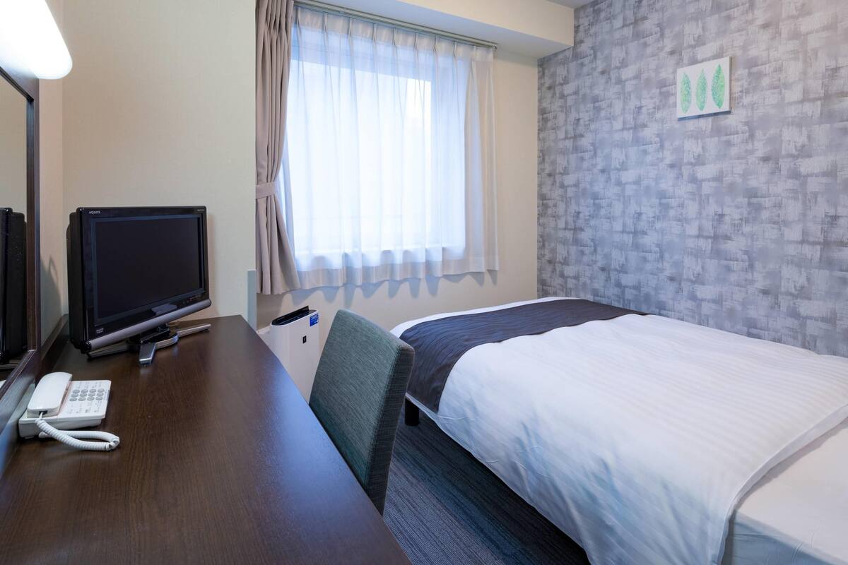 A hotel room with a bed, a desk with a computer monitor, a telephone, and a window with curtains.