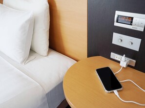 Una habitación de hotel con cama, una mesita de noche con un teléfono cargando y un termostato digital.