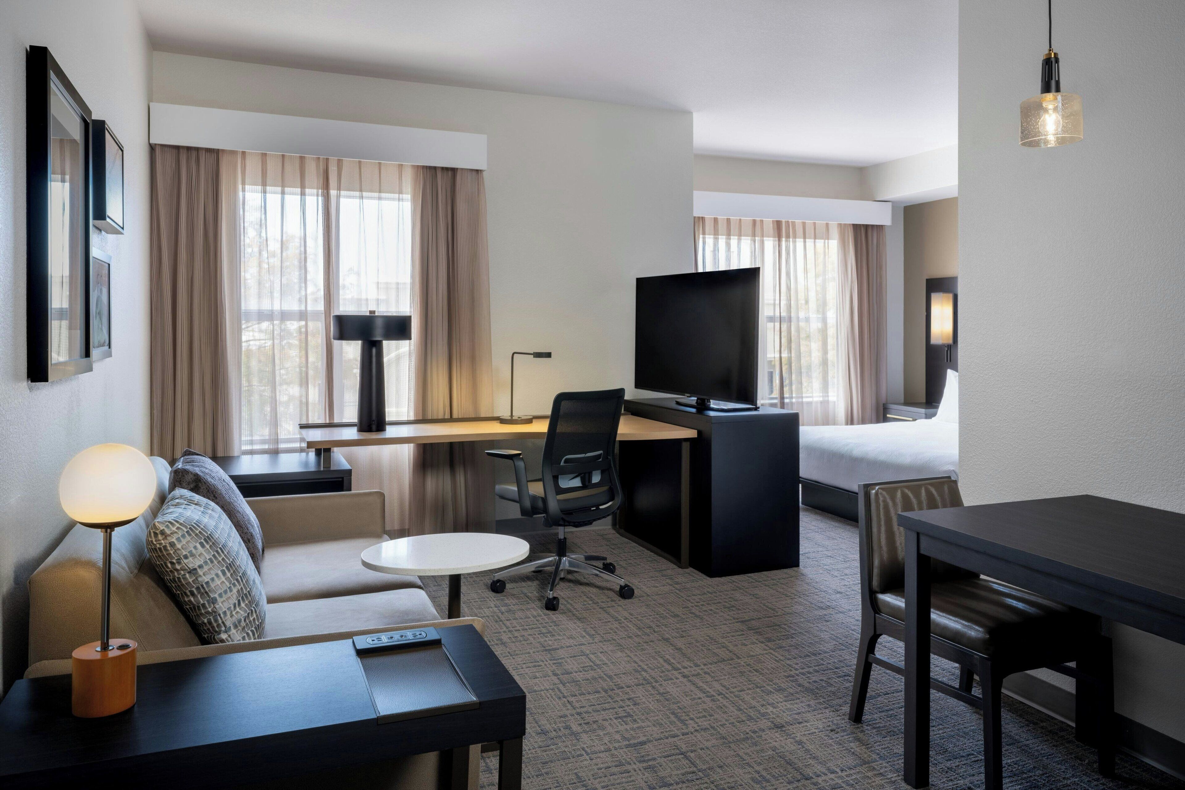 A hotel room with a bed, a desk, a chair, a sofa, a TV, and a lamp.