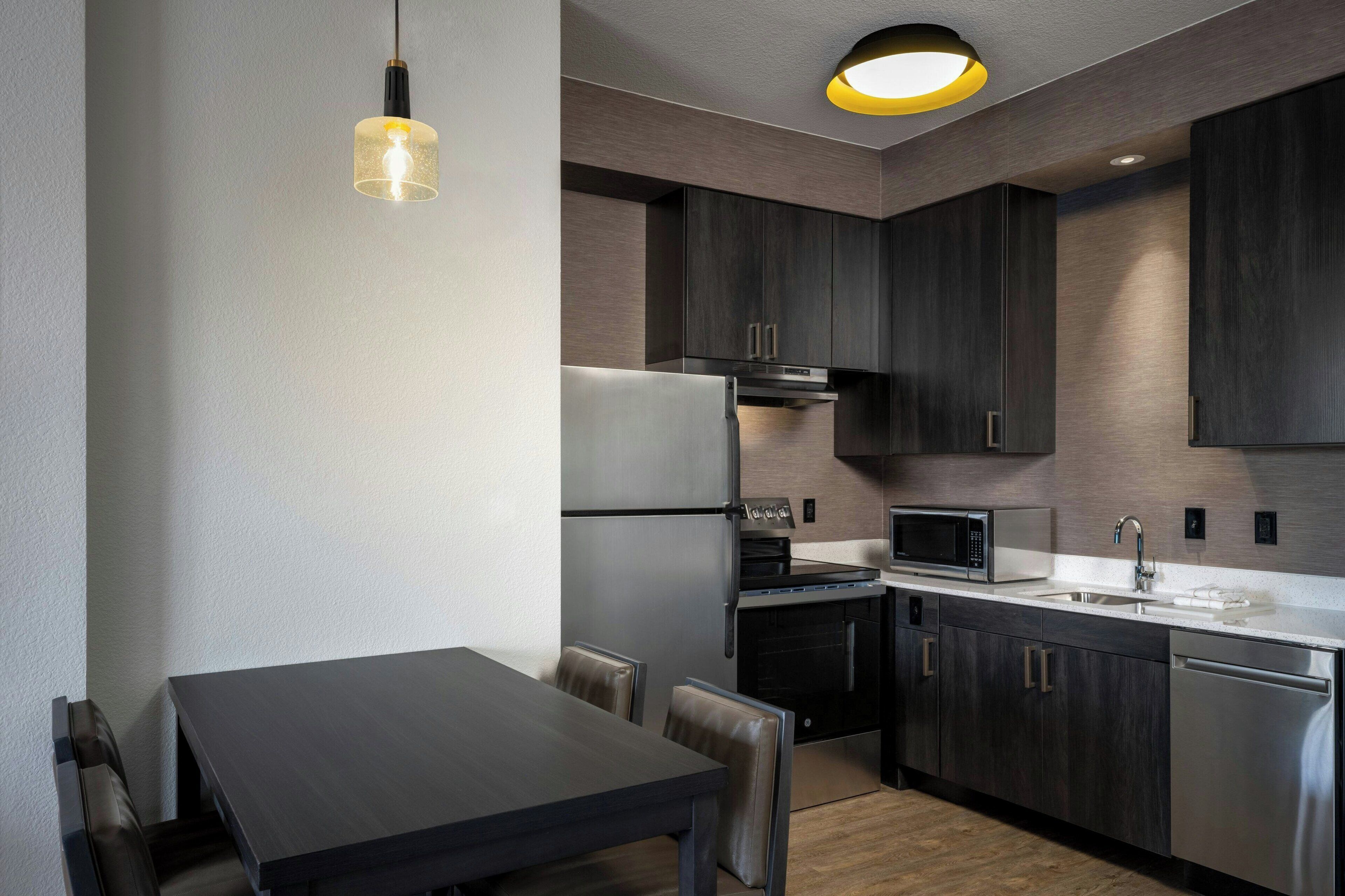 A modern kitchen with dark cabinetry, a stainless steel refrigerator, a microwave, and a built-in oven. There is a dining table with chairs and a pendant light above it.