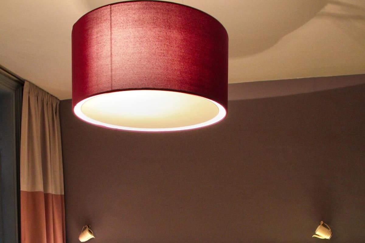 Uma cama de casal com cabeceira escura, duas luminárias de mesa e um pendente cilíndrico vermelho.