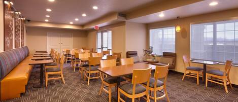 A hotel dining area with multiple tables and chairs, a long bench, and large windows providing views.