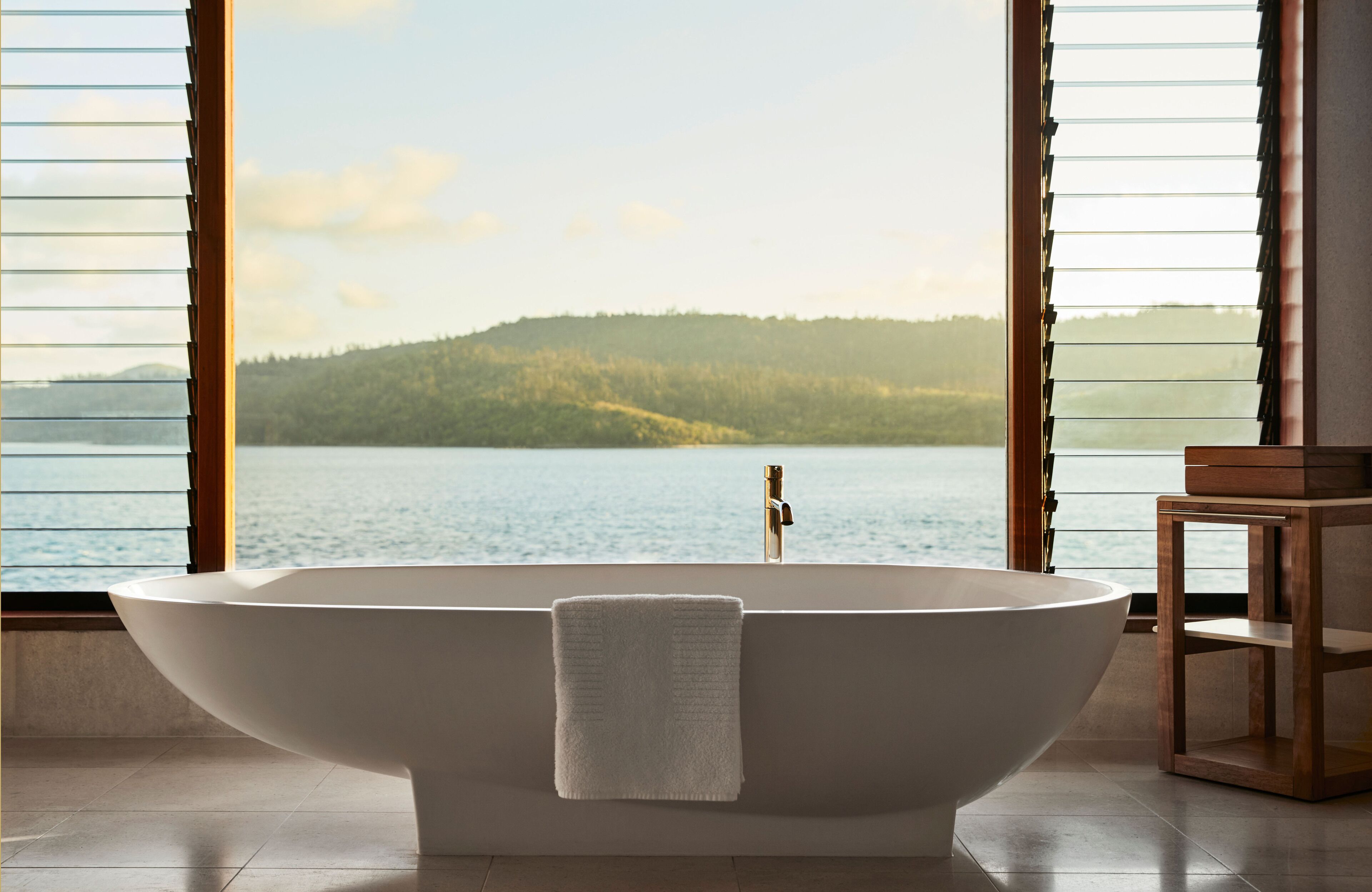Une salle de bain moderne avec une baignoire sur pieds, une table d’appoint en bois et une vue sur un lac et une forêt, grâce à de grandes fenêtres.