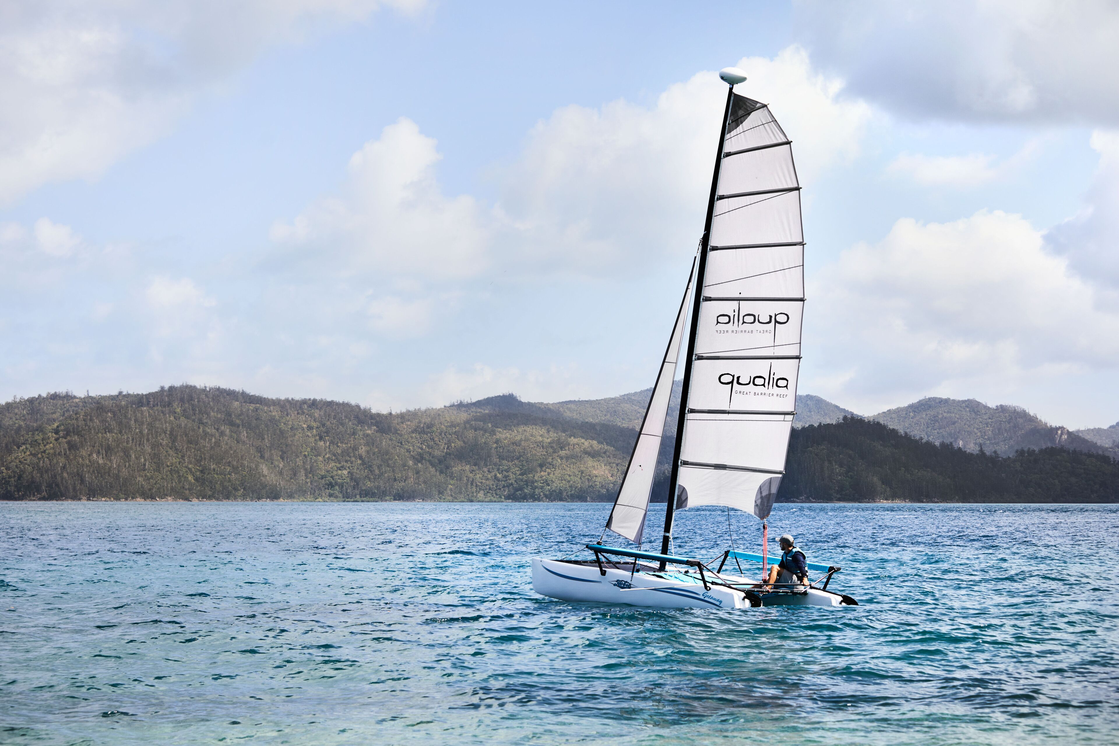 A sailboat with a person on board, sailing on a body of water with a forested hill in the background.