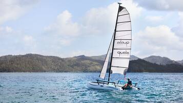 A sailboat with a person on board, sailing on a body of water with a forested hill in the background.