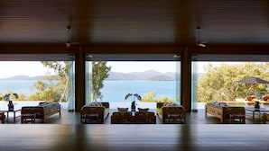 A spacious room with a large window overlooking a body of water and mountains. There are several sofas and a dining table in the center. - Qualia (Whitsundays)