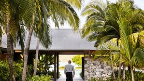 A man walking on a concrete path towards a building with a stone wall and a metal roof. - Qualia (Whitsundays)