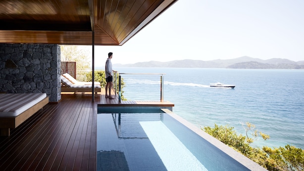 A modern outdoor pool area with a stone wall, wooden deck, and a view of the sea. - Qualia (Whitsundays)