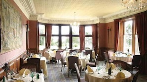 A dining room with multiple tables set for service, large windows with curtains, and a chandelier.