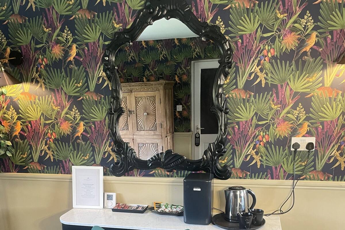 A room with a desk, a teal chair, a decorative mirror, and a patterned wallpaper.