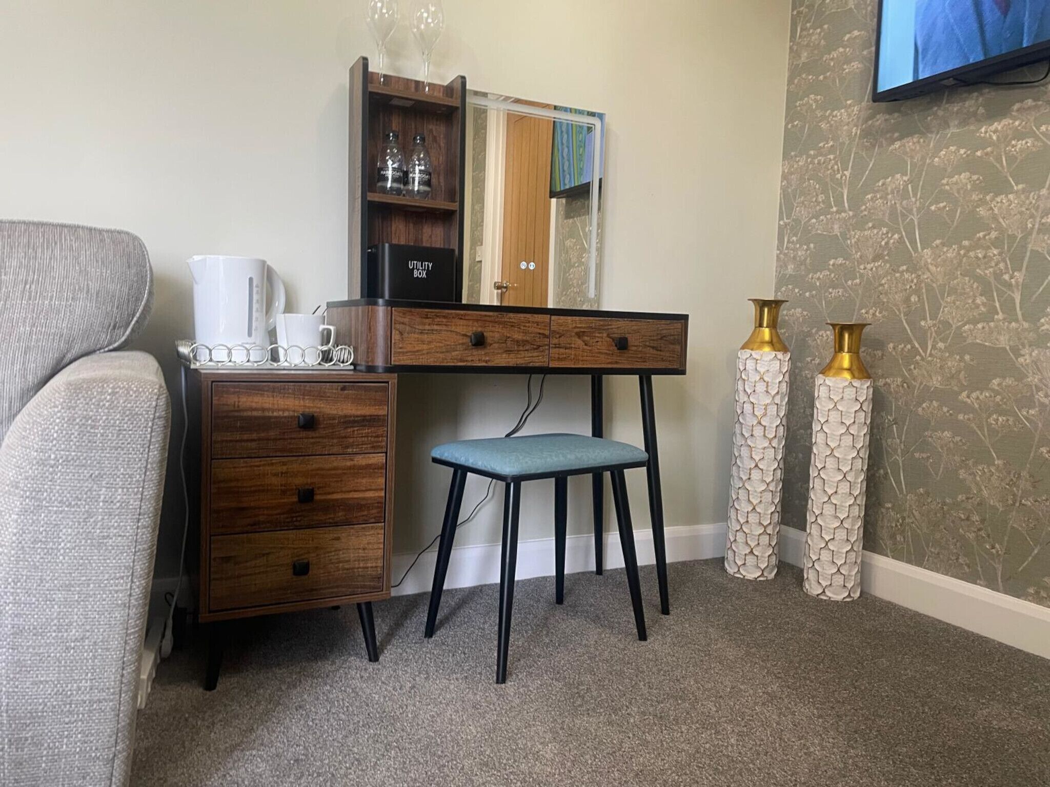 Habitación de hotel compacta con un escritorio de madera, una pequeña silla azul, una cafetera blanca y dos jarrones decorativos.