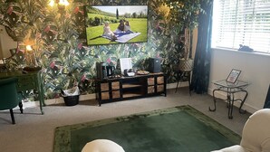 A living room with a large TV mounted on a wall with a tropical wallpaper, a green sofa, a small table, and a coffee table. - The Swan Motel (Beccles)