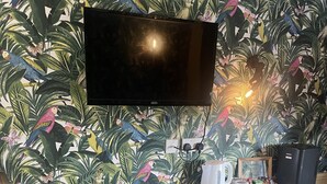 A room with a television mounted on the wall, a wooden bed, a small shelf with a white mug and a box labeled "TEA", and a trash bin. - The Swan Motel (Beccles)