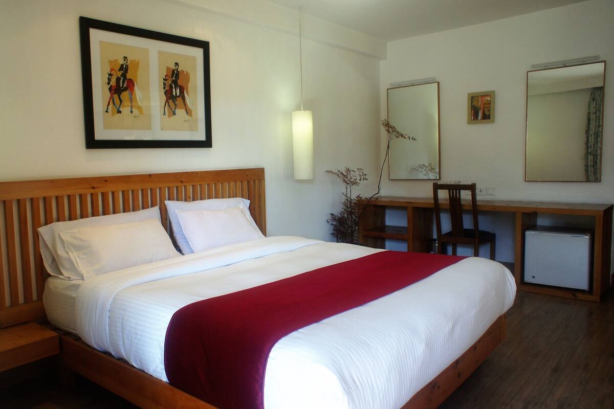 A hotel room with a wooden bed, white linens, a red throw blanket, a desk with a chair, a small refrigerator, and two framed pictures on the wall.