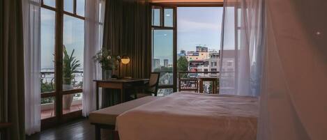 A hotel room with a large bed, a desk with a lamp, and a view of the city through large windows.