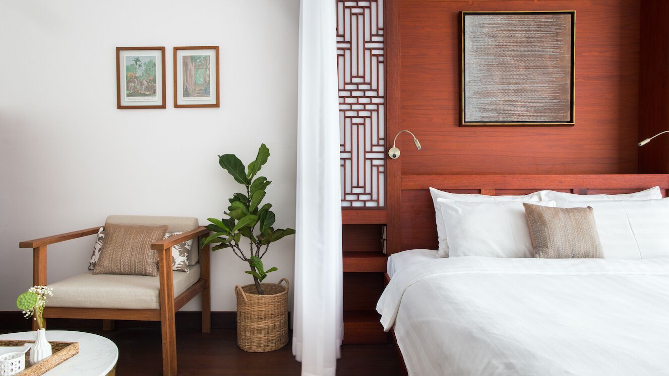 A hotel room with a bed, a chair, a plant, a wall with framed pictures, and a decorative screen.