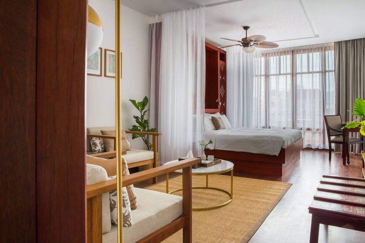 Ein Hotelzimmer mit Bett, Schreibtisch, Stuhl, Sofa, Deckenventilator und einem großen Fenster mit Vorhängen.