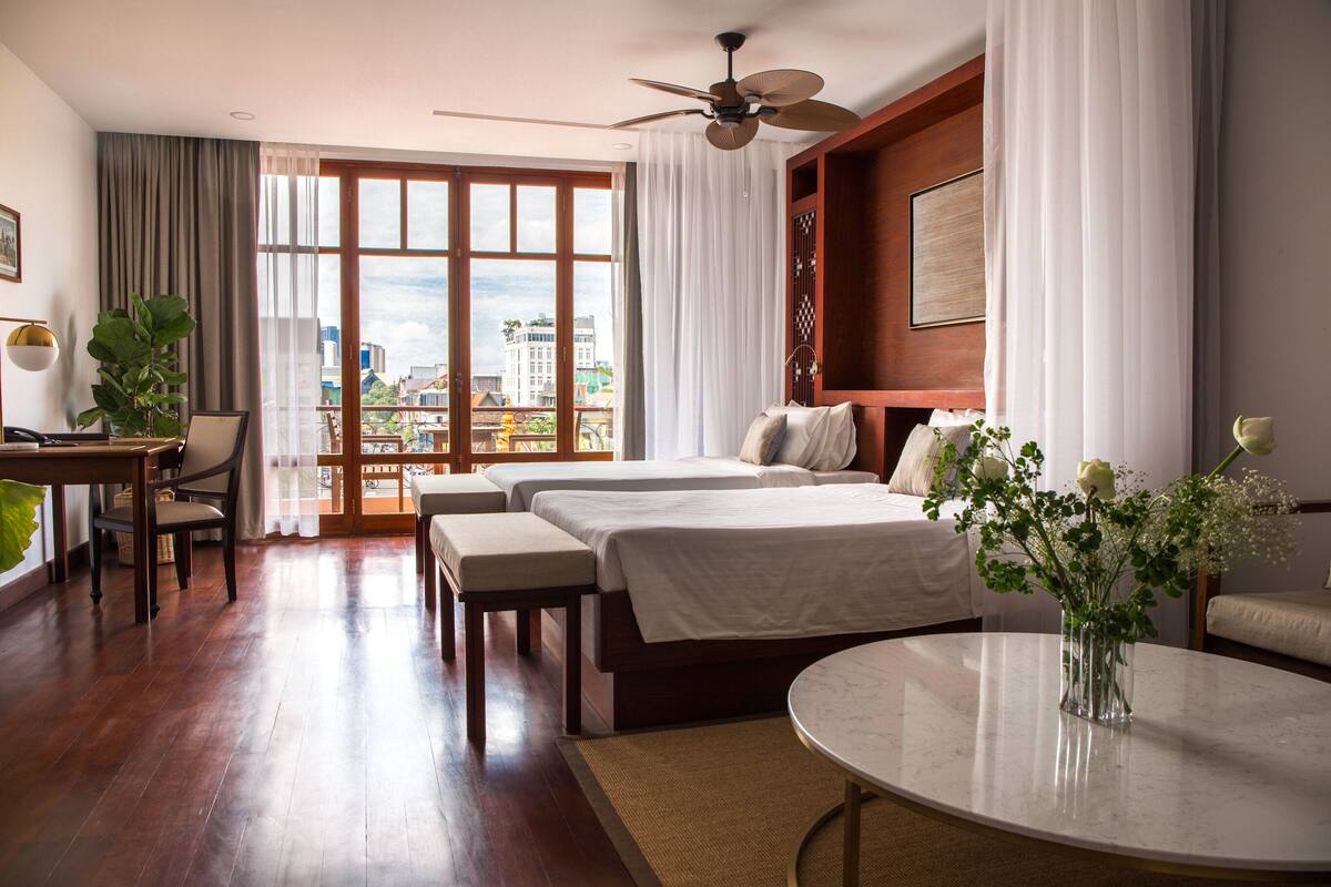 Ein Hotelzimmer mit zwei Betten, einem Esstisch, einem Stuhl, einem runden Couchtisch und Blick auf die Stadt durch große Fenster.