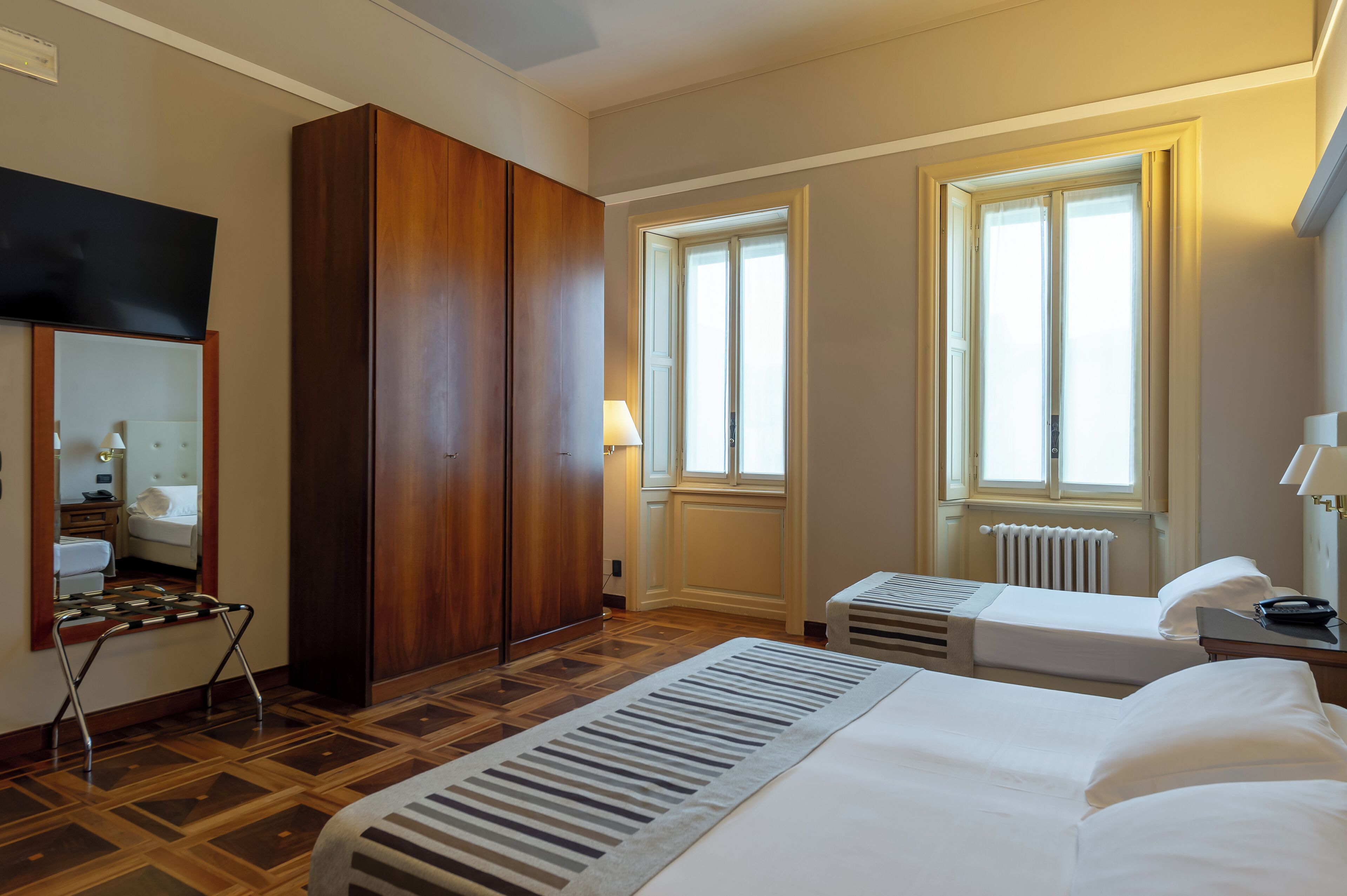 A hotel room with a large bed, two bedside tables, a wardrobe, a mirror, and two windows.