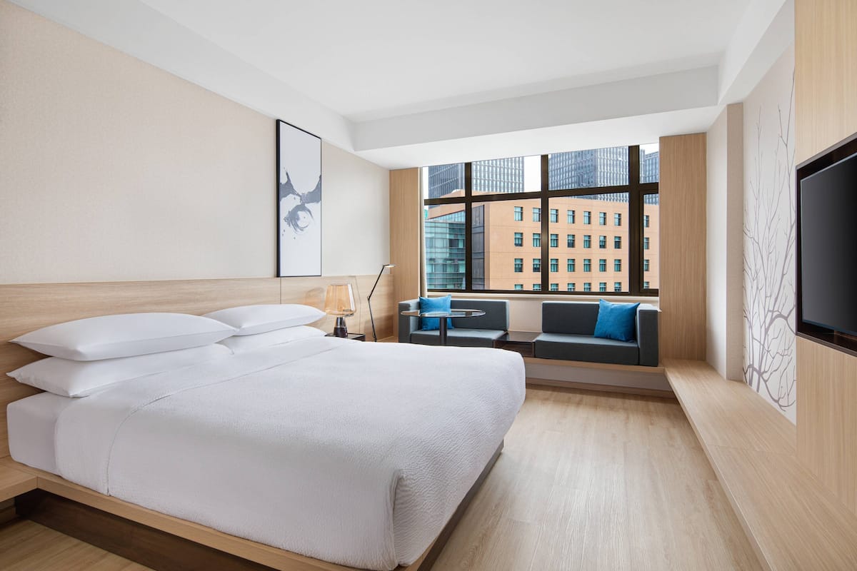 Une chambre d’hôtel moderne dotée d’un grand lit, d’un coin salon avec vue sur la ville et d’un téléviseur à écran plat fixé au mur.