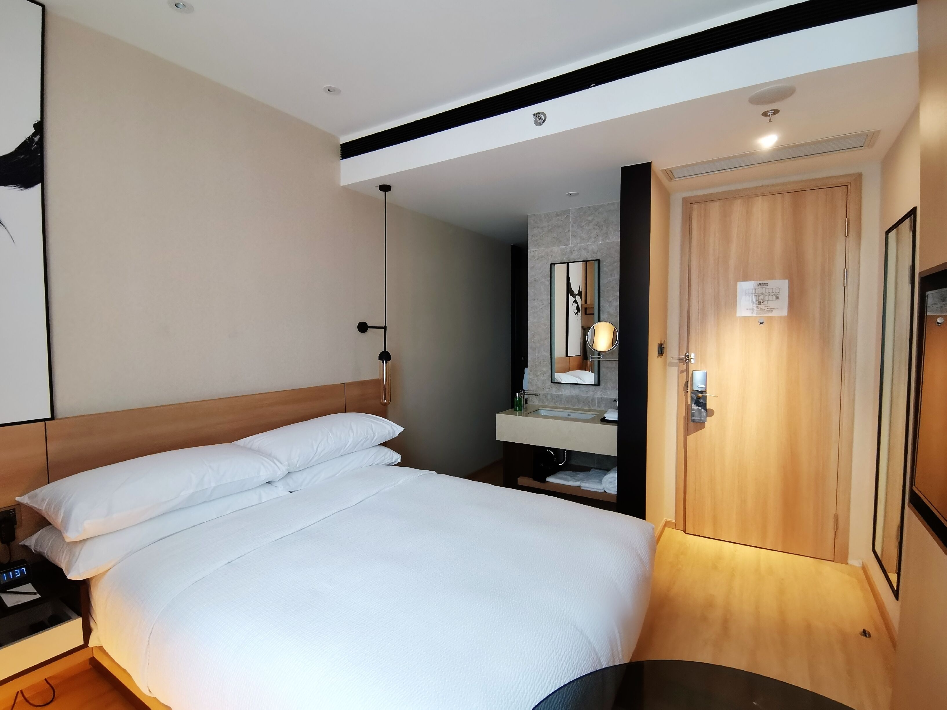 A hotel room with a large bed, a bedside table with a lamp, a mirror above the sink, and a wooden door.