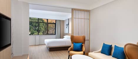 A modern hotel room with a bed, a sofa, a chair, a TV, and a large window offering a view of trees and buildings.