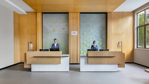 Zona de recepción con dos recepcionistas en mostradores, paneles de madera, un ventanal y una pared con notas.