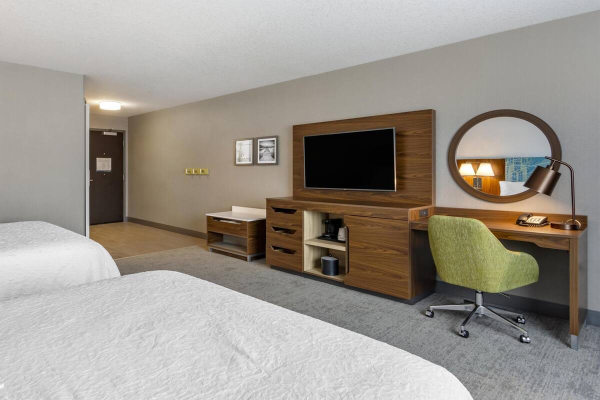 A hotel room with two beds, a wooden desk, a green office chair, a flat-screen TV, a round mirror, and a small shelf with a lamp.
