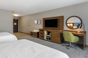 Une chambre d’hôtel avec deux lits, un bureau en bois, une chaise de bureau verte, un téléviseur à écran plat, un miroir rond et une petite étagère avec une lampe.