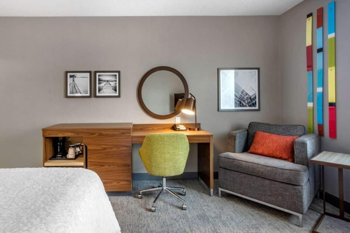 A hotel room with a bed, a desk with a chair, a round mirror, and two framed pictures on the wall.