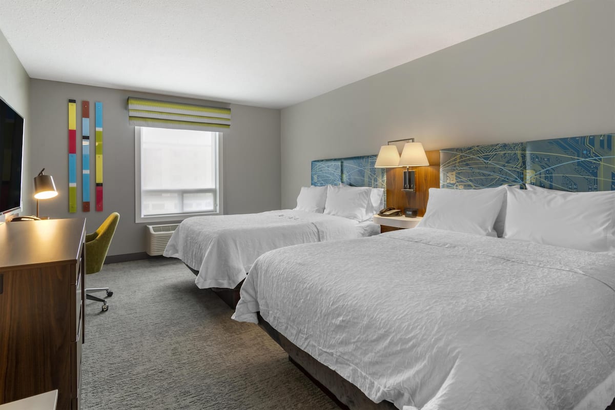 A hotel room with two beds, a desk, a chair, a lamp, and a window.