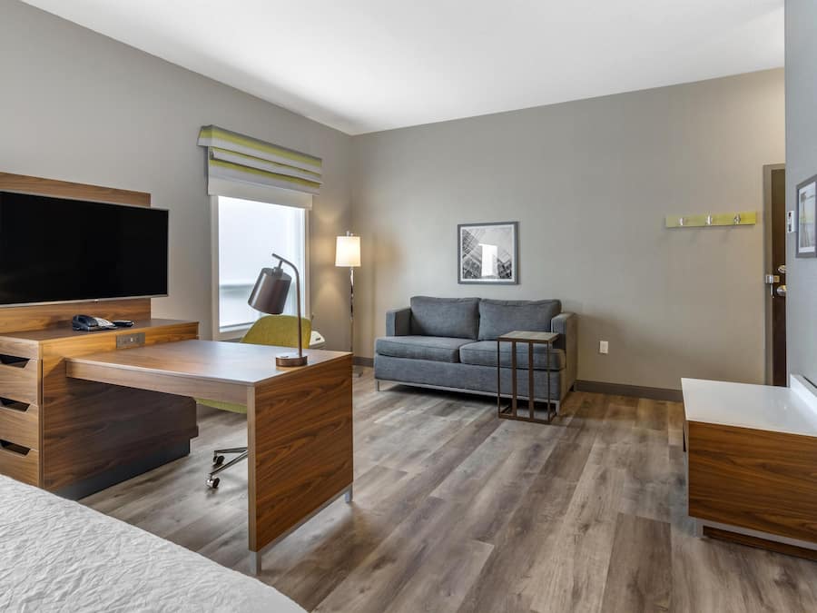 Une chambre d’hôtel moderne avec un bureau en bois, un canapé gris et un téléviseur à écran plat.