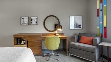 A hotel room with a wooden desk, a green office chair, a grey sofa, a round mirror, and framed pictures on the wall.