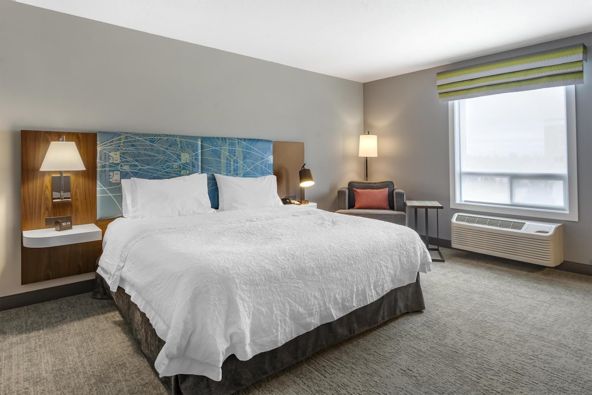 A hotel room with a large bed, bedside tables with lamps, a chair, a window with blinds, and a wall-mounted air conditioner.