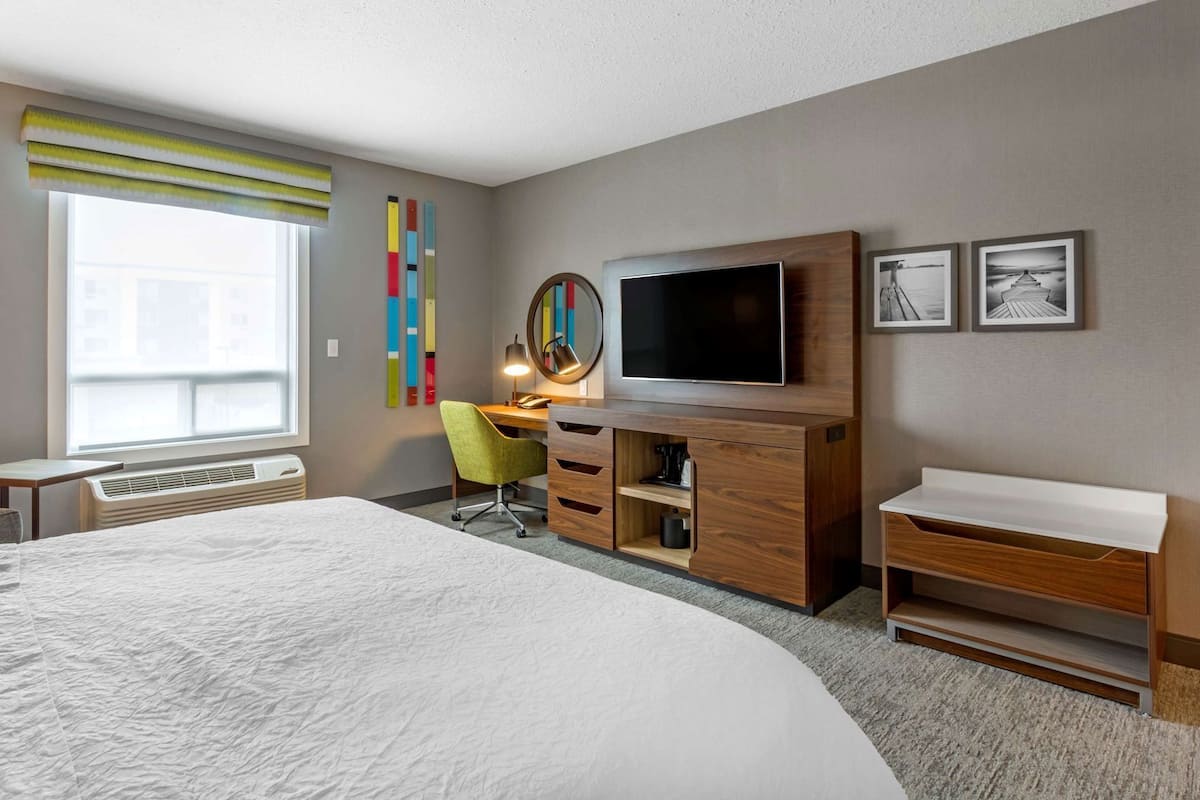 A hotel room with a large bed, a desk with a TV, a chair, a mirror, and artwork on the wall.