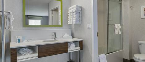 Un baño moderno con lavamanos, espejo y retrete blancos. Cuenta con un box de ducha de vidrio y un perchero con toallas dobladas.
