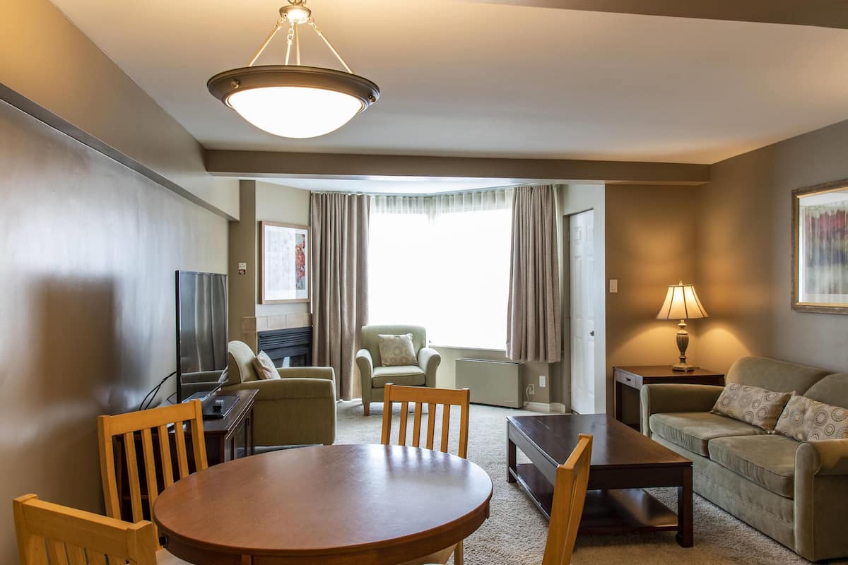 Hotelkamer met een eettafel, stoelen, een bank, een televisie, een lamp en een groot raam met gordijnen.