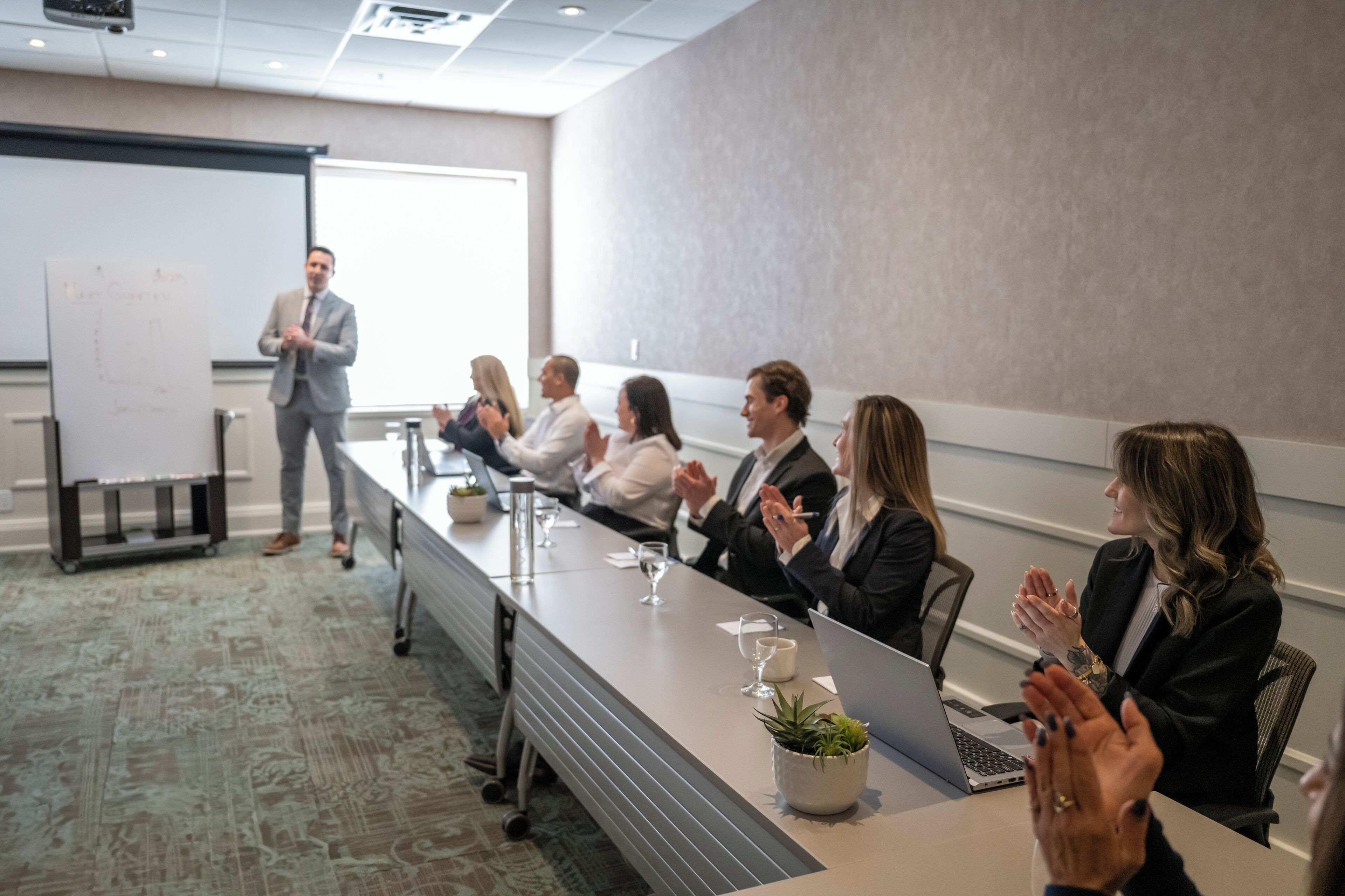 A conference room with a long table, a projector screen, and attendees clapping.