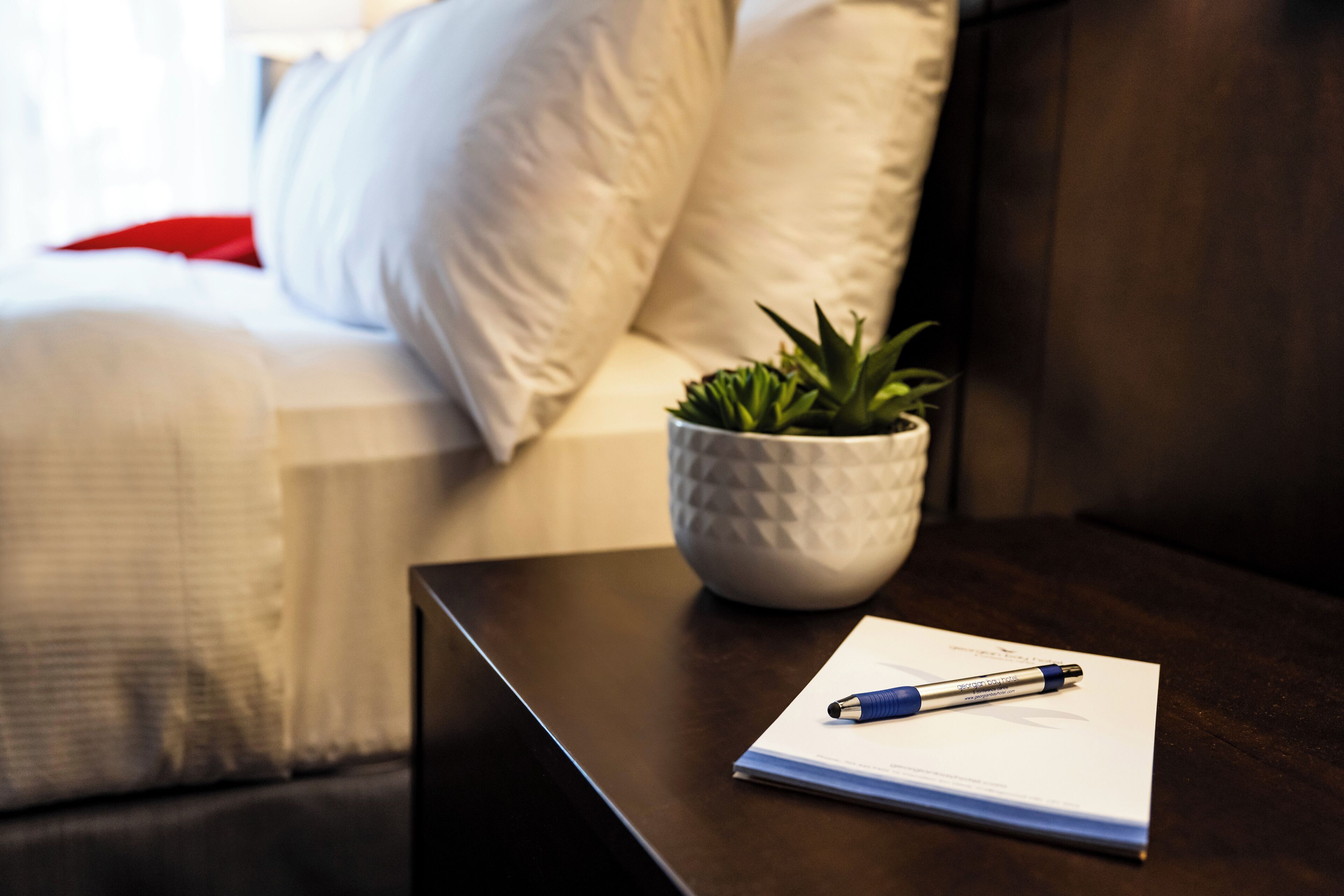 Quarto de hotel bem arrumado com uma cama, uma mesinha de cabeceira com um caderno e caneta, e um vaso de planta.