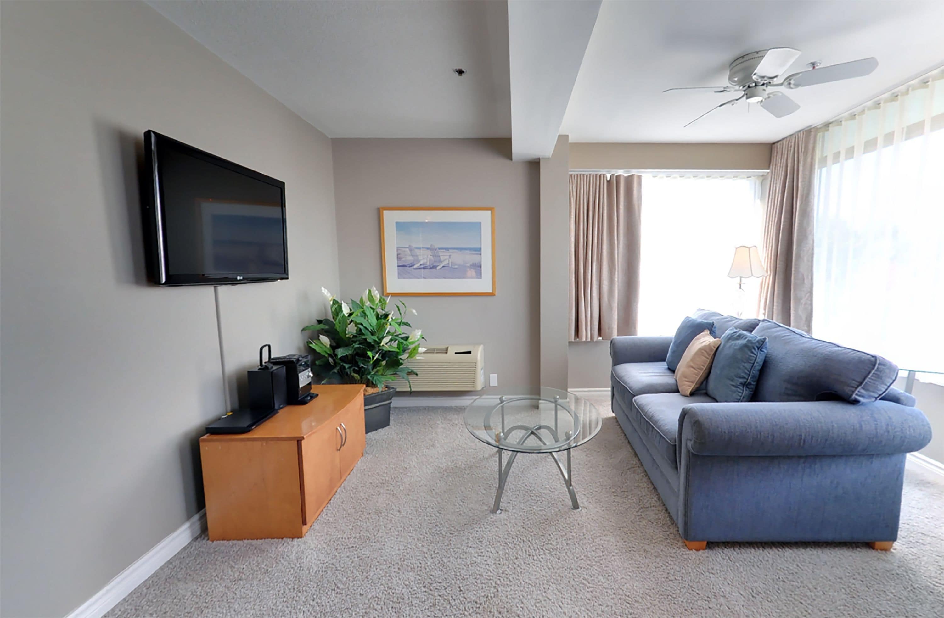 A living room with a flat-screen TV, a blue sofa, a glass coffee table, a wooden cabinet, a potted plant, and a ceiling fan.
